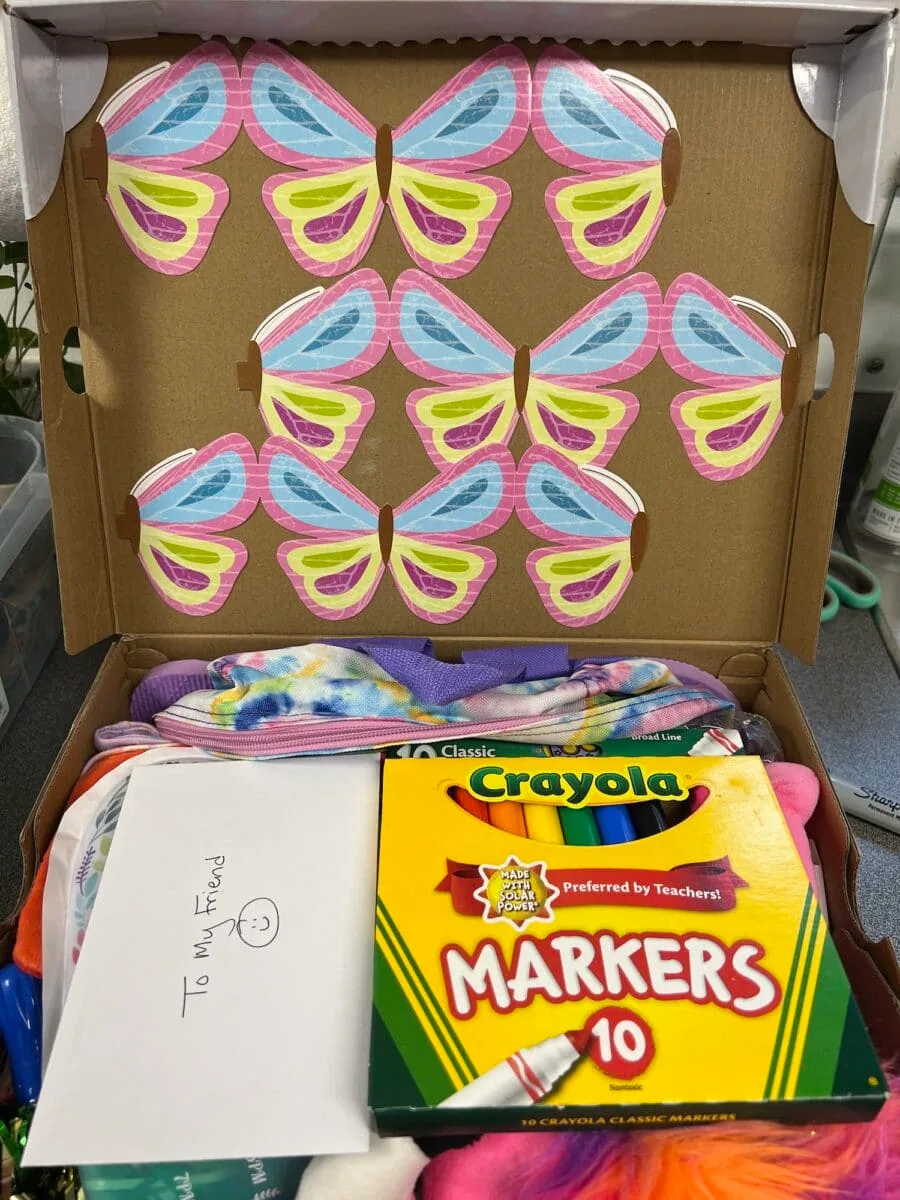 An open box decorated with colorful butterfly cutouts, filled with Crayola markers, an envelope labeled "To My Friend :)", and other small gifts—perfect for an Operation Christmas Child donation.