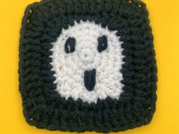 A ghost granny square with a black border and a white center featuring a black smiling face, placed on a yellow background.