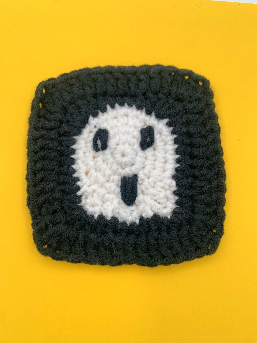 A ghost granny square with a black border and a white center featuring a black smiling face, placed on a yellow background.