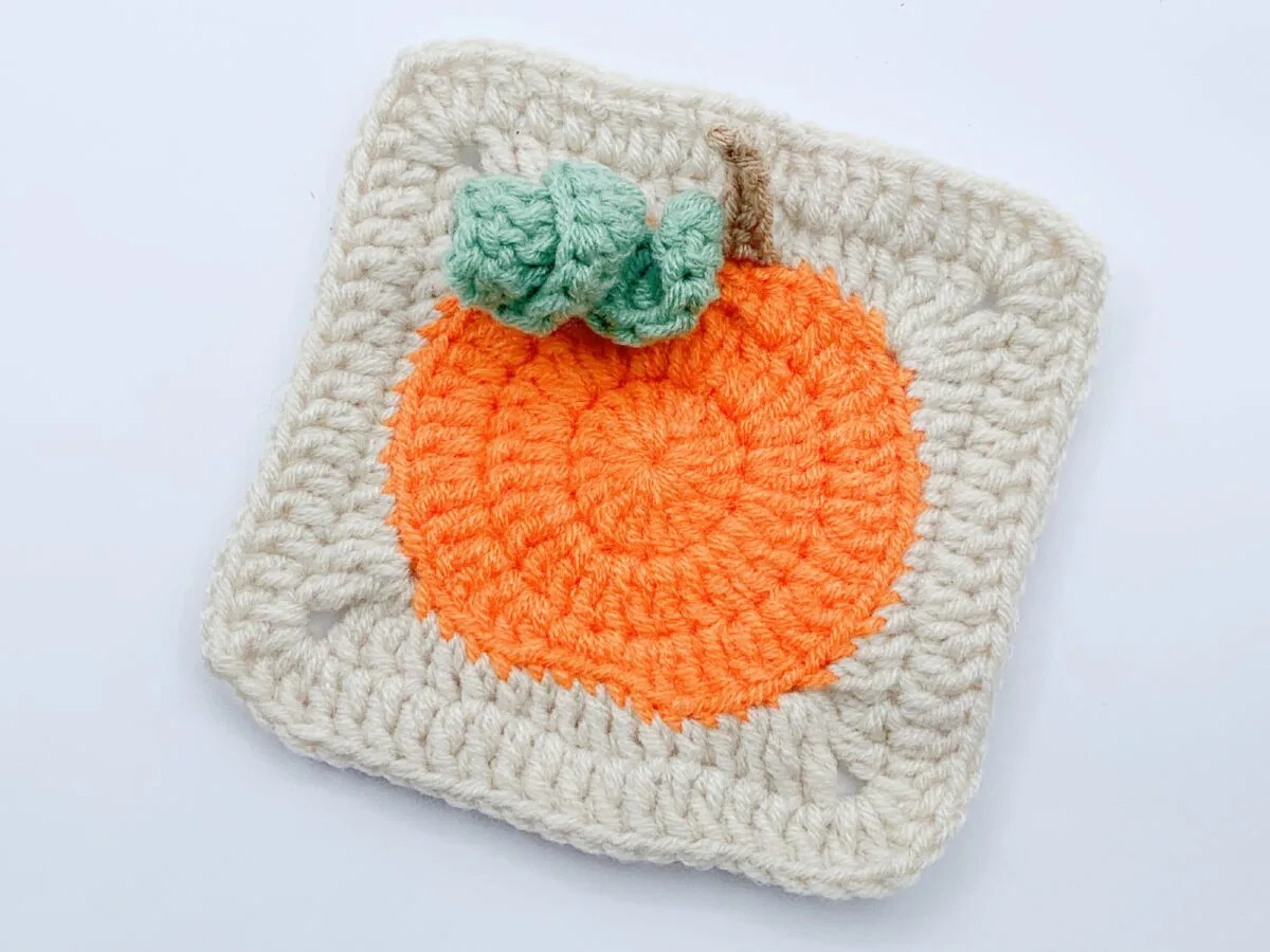 A crocheted square featuring an orange circle with a green leaf and brown stem, resembling a pumpkin, on a white background.