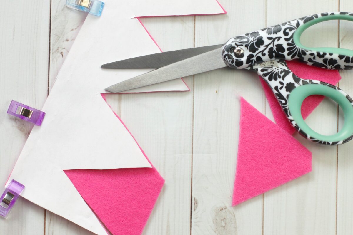 A pair of patterned scissors, pink felt triangles, white paper, and small clips on a light wooden surface.