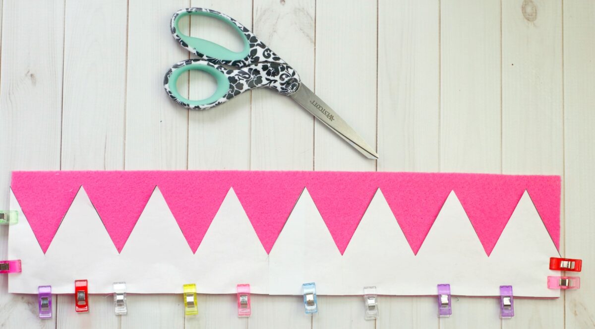 A pair of patterned scissors and a strip of pink felt with white zigzag paper pinned by colorful clips on a light wooden surface.