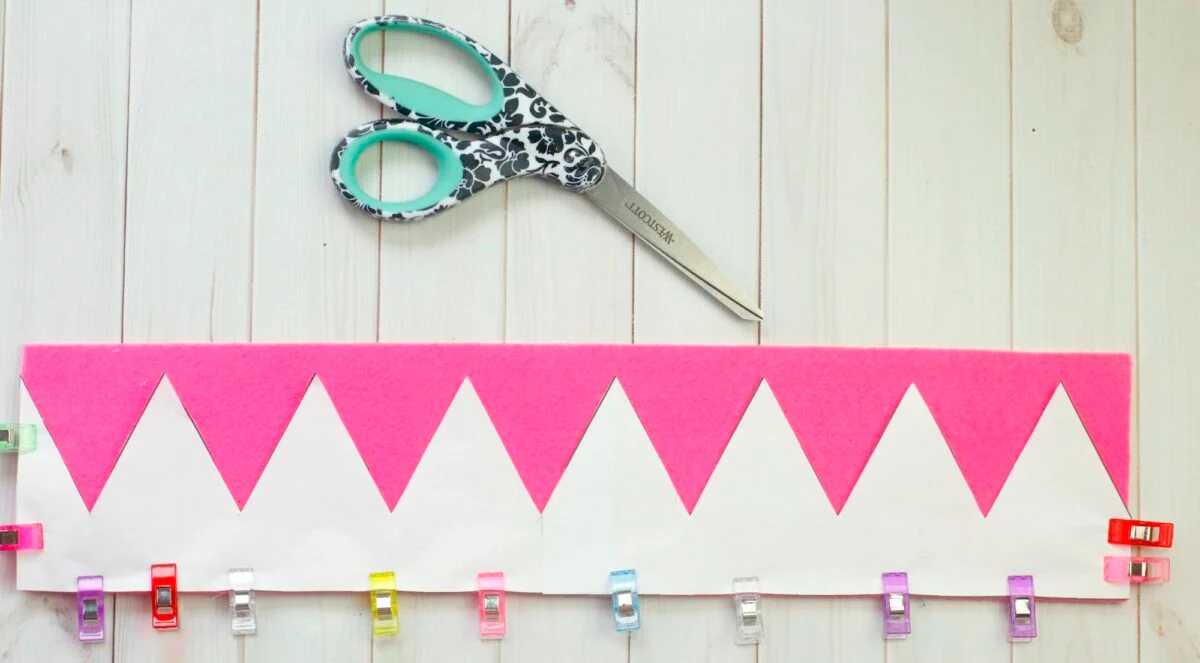 A pair of patterned scissors and a strip of pink felt with white zigzag paper pinned by colorful clips on a light wooden surface.