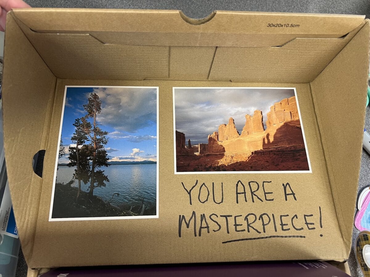 Two landscape photos in a cardboard box: one of a lake with trees, the other of red rock formations. "YOU ARE A MASTERPIECE!" is handwritten below, echoing the heartfelt spirit behind every Operation Christmas Child gift.