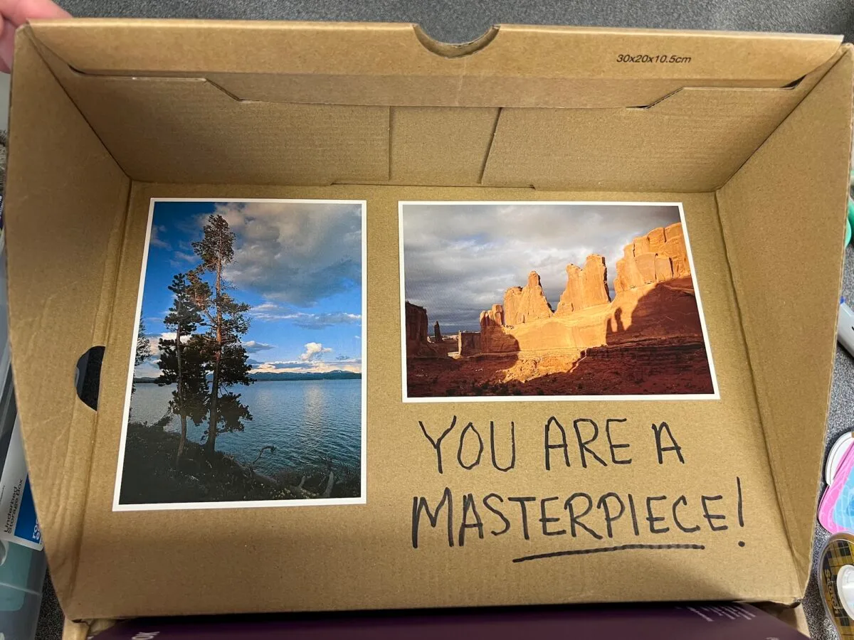 Two landscape photos in a cardboard box: one of a lake with trees, the other of red rock formations. "YOU ARE A MASTERPIECE!" is handwritten below, echoing the heartfelt spirit behind every Operation Christmas Child gift.