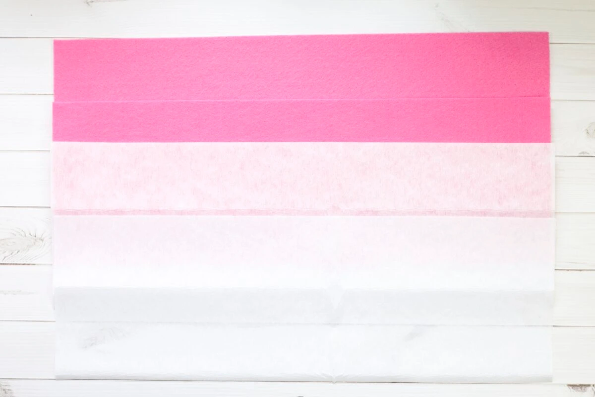 Four large sheets of tissue paper in descending shades from bright pink at the top to white at the bottom, laid out on a white wooden surface.