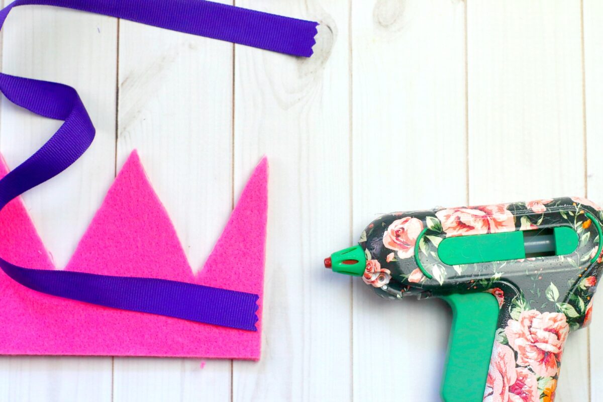 A pink felt crown, a purple ribbon, and a floral-patterned hot glue gun are placed on a white wooden surface.