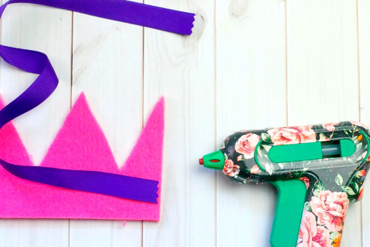 A pink felt crown, a purple ribbon, and a floral-patterned hot glue gun are placed on a white wooden surface.