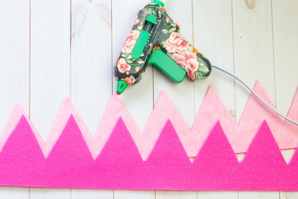 A floral-patterned glue gun is placed on a wooden surface next to a pink felt zigzag strip, suggesting a craft project in progress.