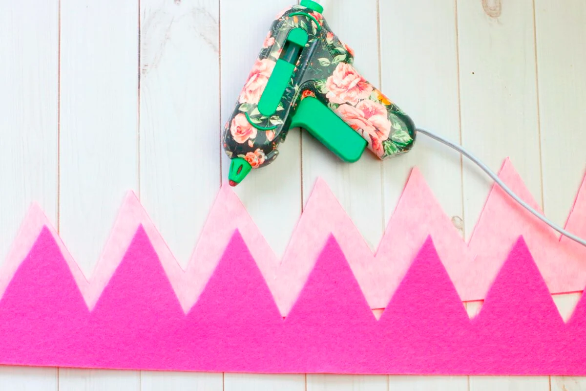 A floral-patterned glue gun is placed on a wooden surface next to a pink felt zigzag strip, suggesting a craft project in progress.