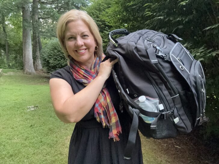 A woman standing outside holds a large black backpack bug out bag over her shoulder, with a water bottle visible in a side pocket.