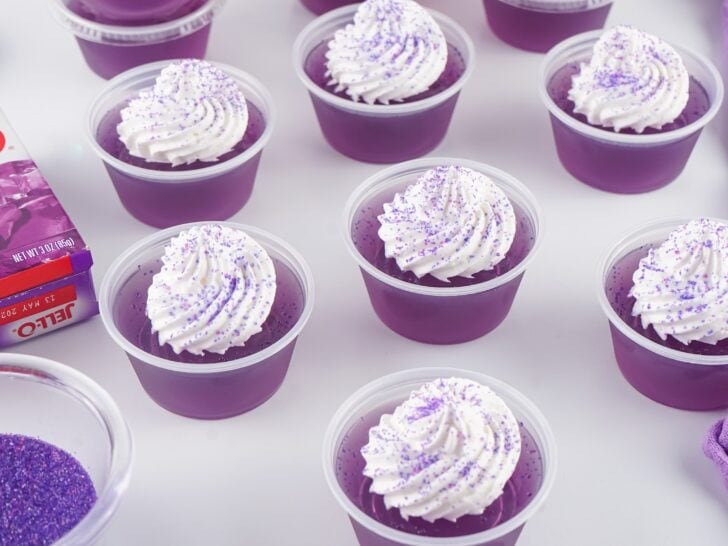 Several small clear cups filled with purple gelatin, each topped with a swirl of whipped cream and purple sprinkles, are arranged on a white surface with a box of Jell-O nearby.