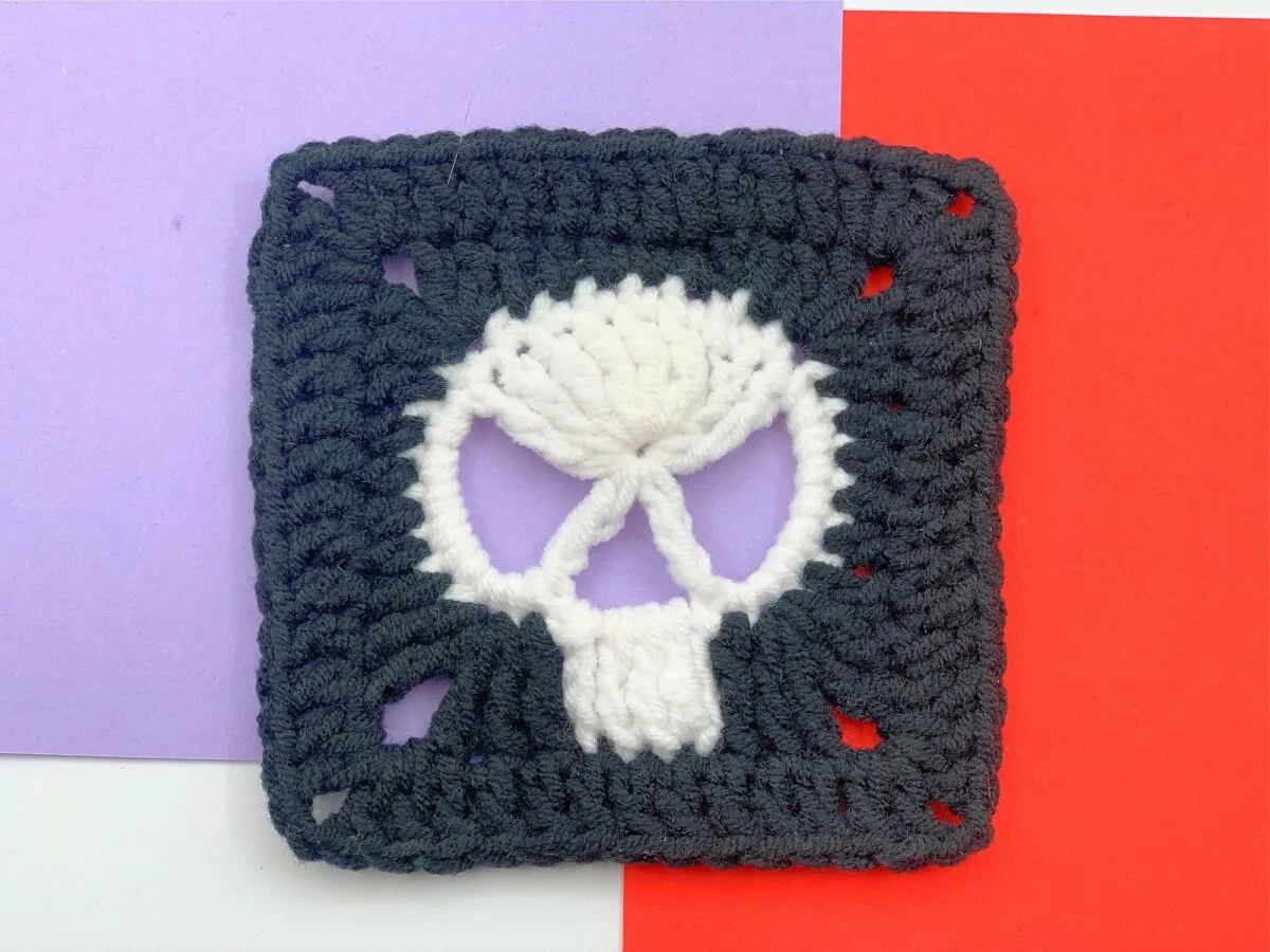 A crocheted black square with a white skull design in the center, placed on a background of purple, red, and white sections.