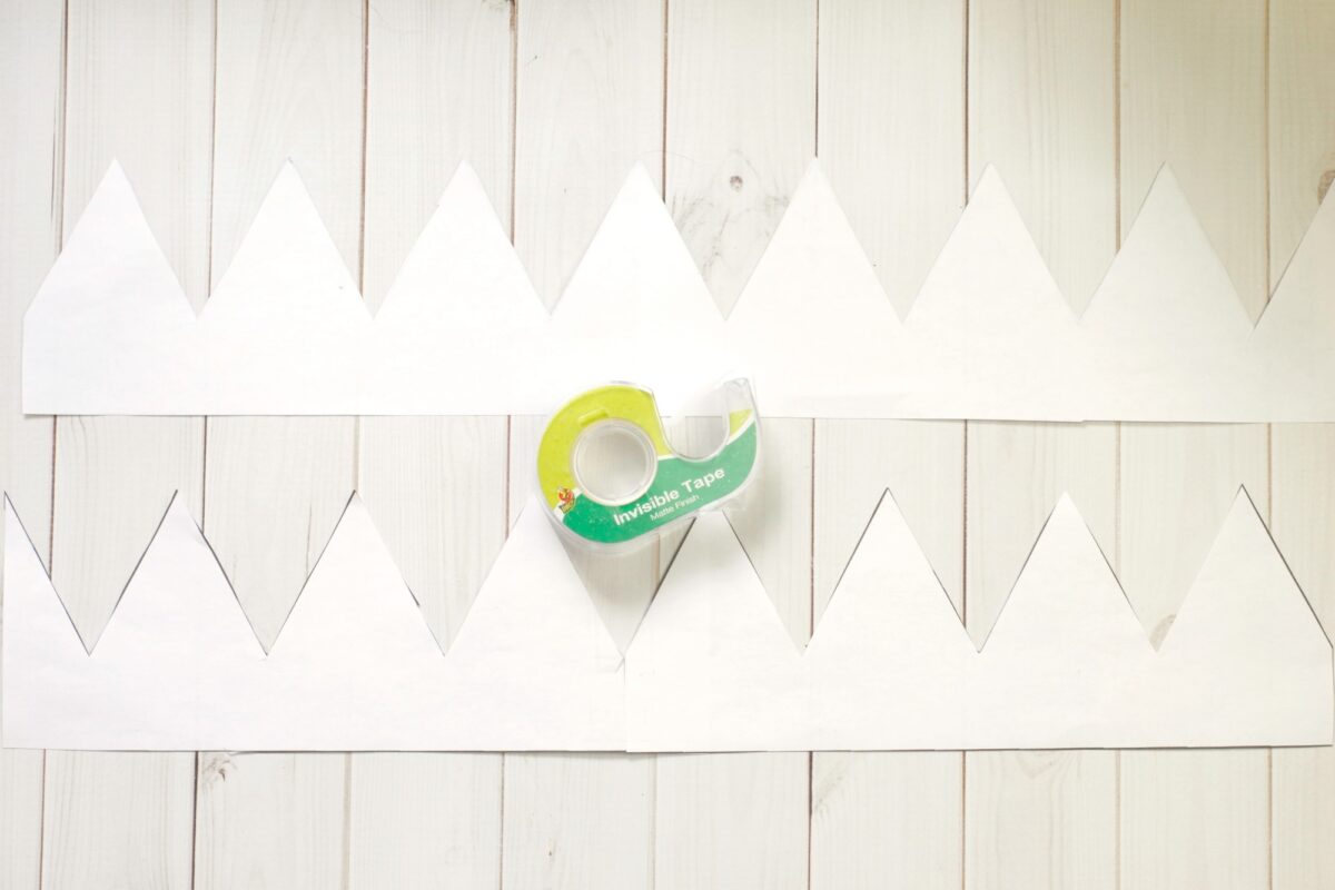 Two strips of white paper cut with pointed edges, resembling crowns, are placed on a wooden surface with a roll of invisible tape in the center.