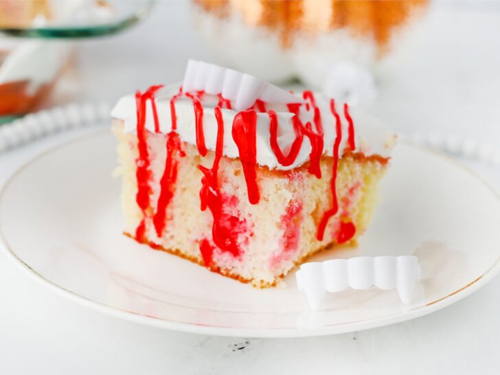 A slice of white cake with red syrup and white frosting on a plate, decorated with plastic vampire fangs beside it.