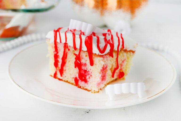 A slice of white cake with red syrup and white frosting on a plate, decorated with plastic vampire fangs beside it.