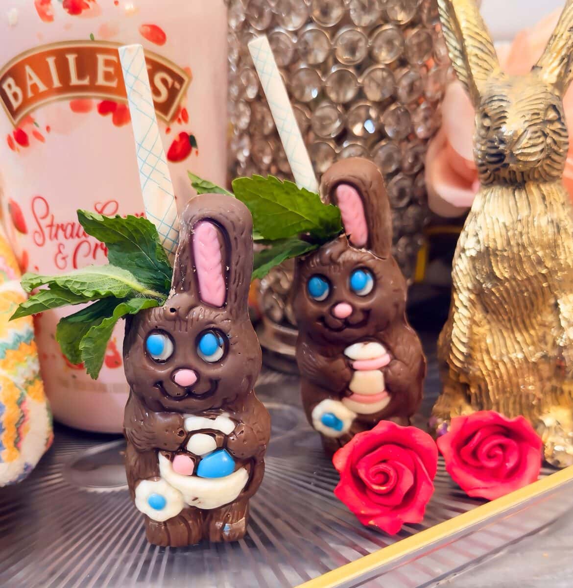 Two chocolate rabbit cocktails with blue eyes, mint garnish, and paper straws sit beside a bottle of Baileys, a gold bunny figurine, and two decorative red roses.