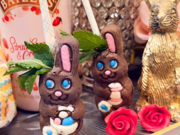 Two chocolate rabbit cocktails with blue eyes, mint garnish, and paper straws sit beside a bottle of Baileys, a gold bunny figurine, and two decorative red roses.