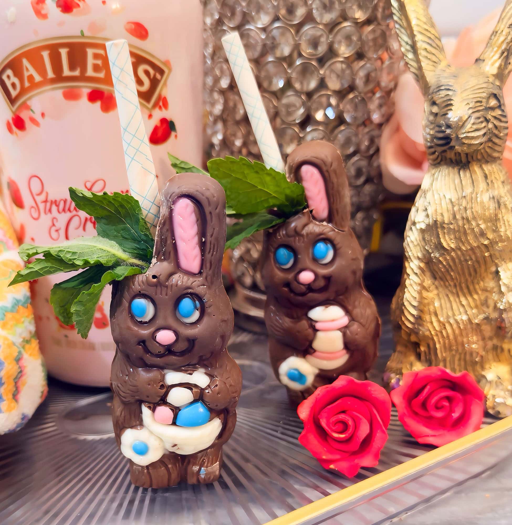 Two chocolate rabbit cocktails with blue eyes, mint garnish, and paper straws sit beside a bottle of Baileys, a gold bunny figurine, and two decorative red roses.