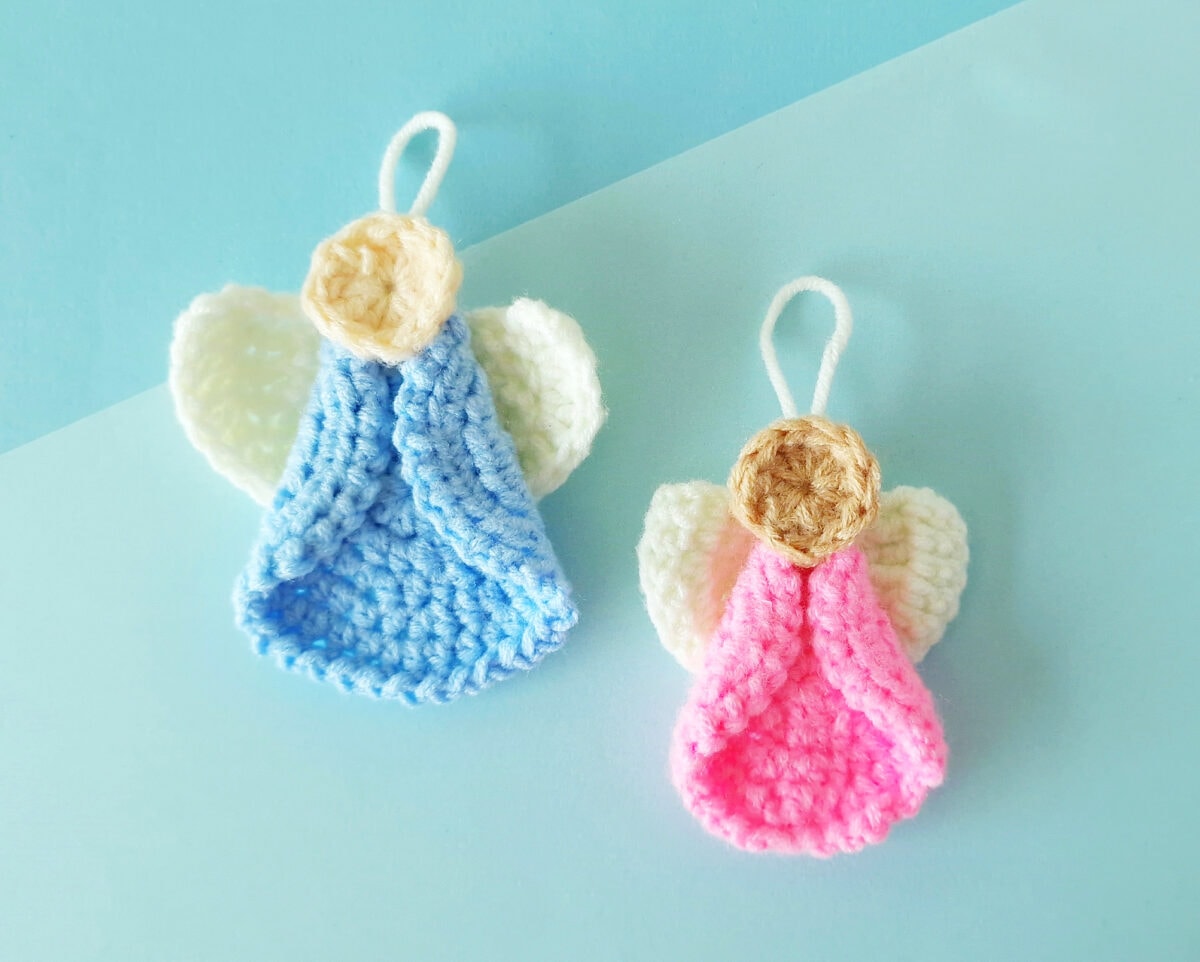 Two handmade crochet angel ornaments with blue and pink bodies, cream-colored wings, and beige heads, displayed on a light blue background.