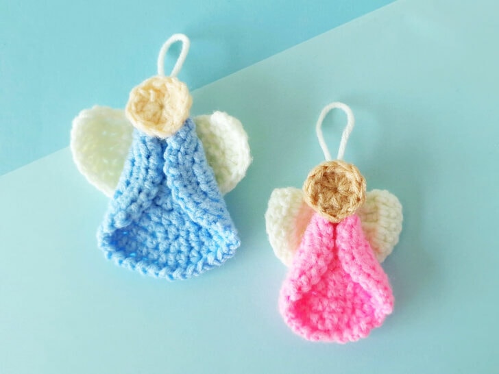 Two handmade crochet angel ornaments with blue and pink bodies, cream-colored wings, and beige heads, displayed on a light blue background.