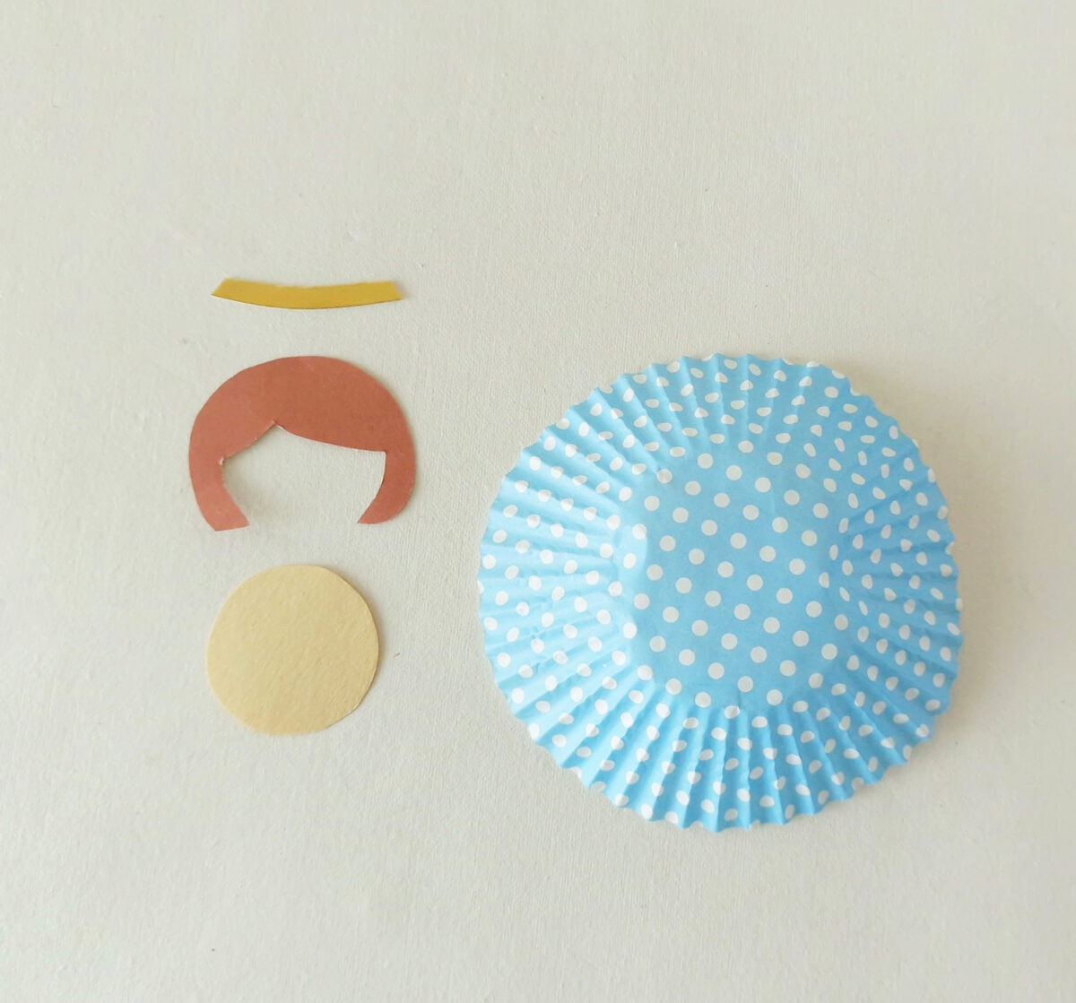 Paper cutouts arranged on a white surface: a blue polka-dot cupcake liner, a round beige piece, a brown hair shape, and a small yellow strip.