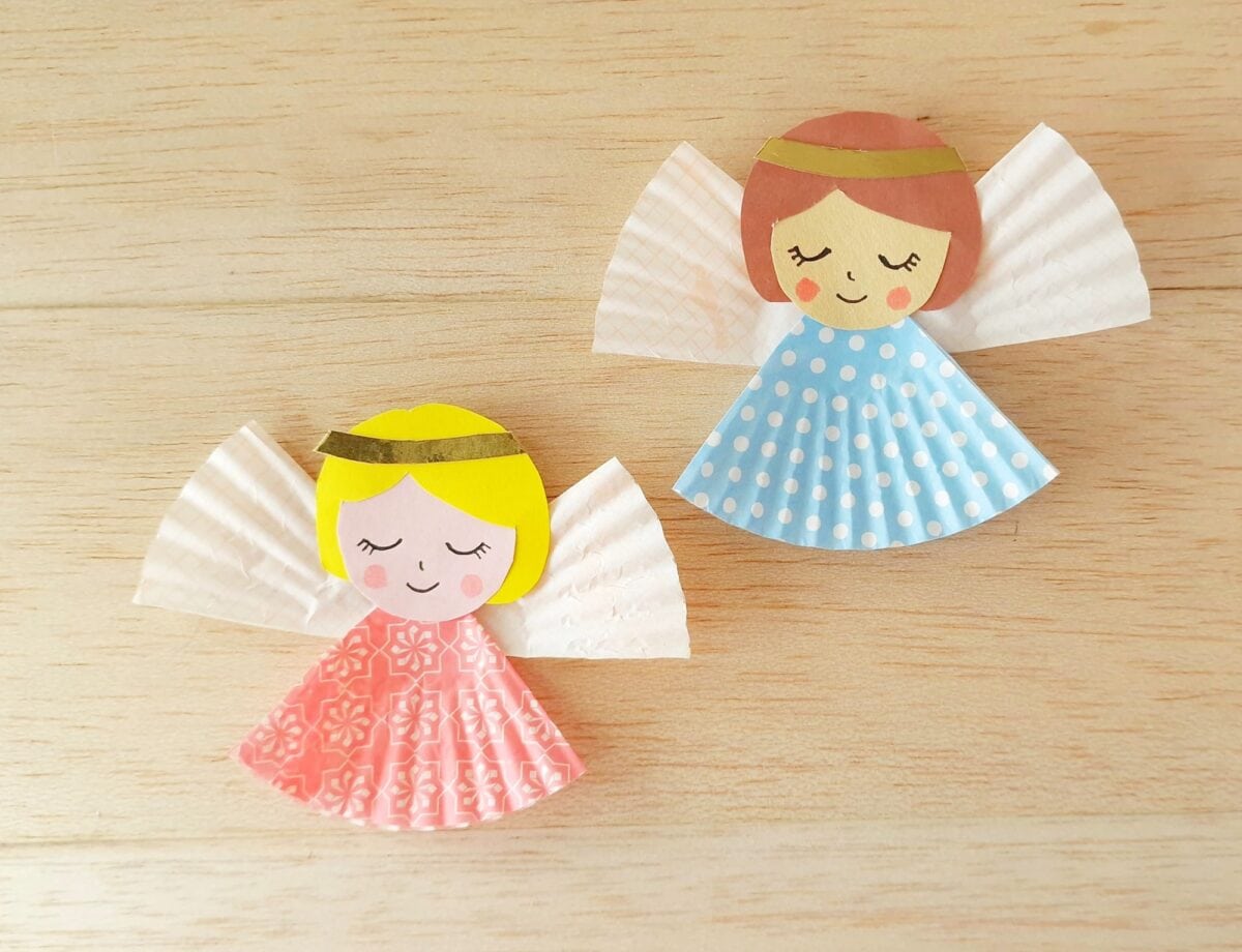 Two handmade paper angel crafts with cupcake liner dresses, one pink and one blue, both with gold headbands, on a light wooden surface.