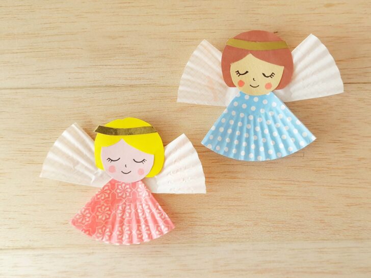 Two handmade paper angel crafts with cupcake liner dresses, one pink and one blue, both with gold headbands, on a light wooden surface.