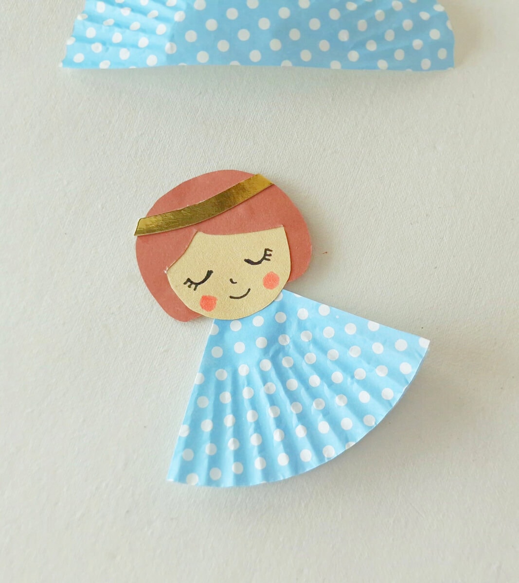 Paper craft angel with a round face, closed eyes, pink cheeks, brown hair, gold headband, and a blue polka dot dress made from a cupcake liner on a white background.