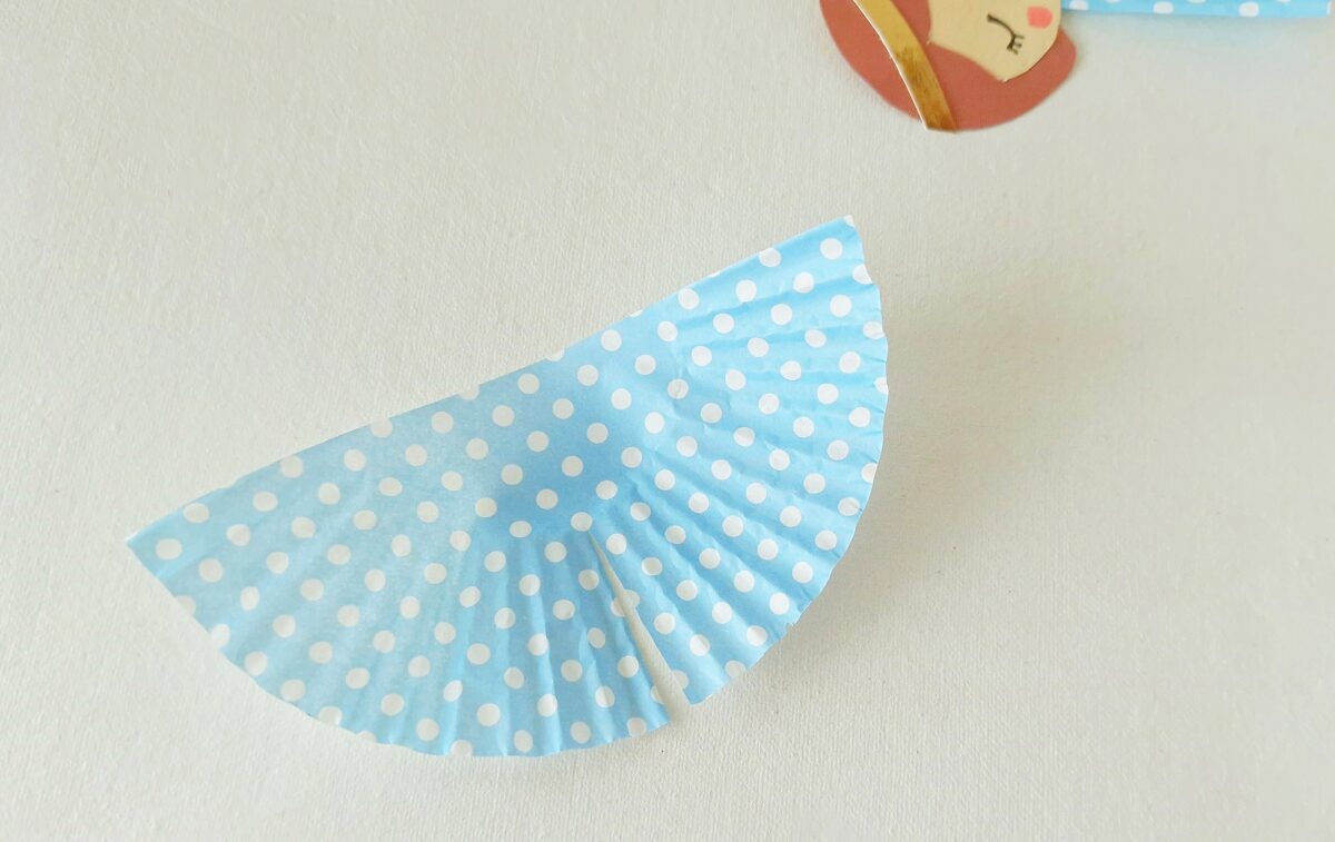 A blue paper cupcake liner with white polka dots is folded in half on a white surface; part of a paper cutout with a face is visible in the upper right corner.