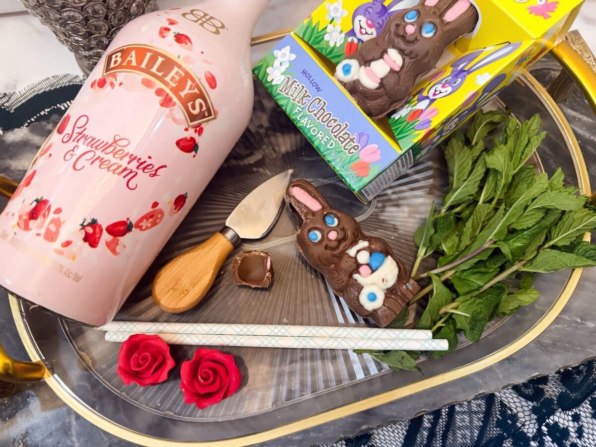 A bottle of Bailey’s Strawberries & Cream, two chocolate bunnies, a cheese knife, mint sprigs, two red rose decorations, and a white straw are arranged on a tray—perfect for crafting a whimsical chocolate rabbit cocktail.