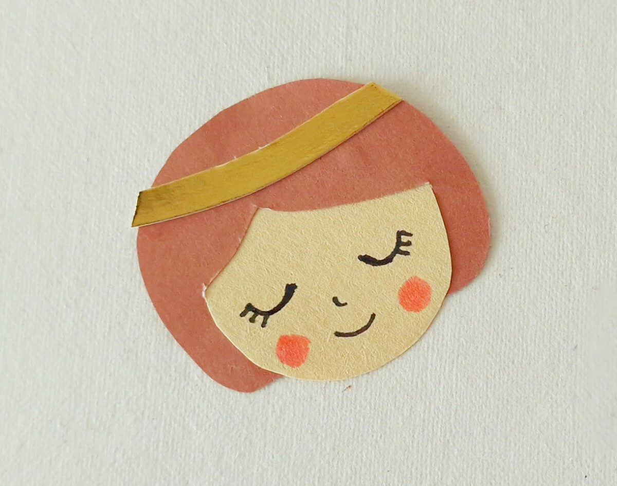 A paper cutout of a smiling face with closed eyes, rosy cheeks, brown hair, and a yellow headband on a light background.