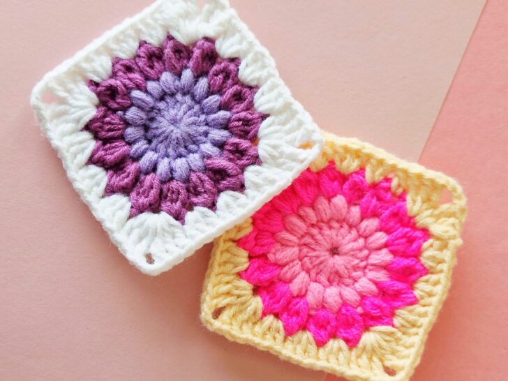 Two colorful crochet sunburst flower granny squares, one with purple and white yarn and the other with pink and yellow yarn, placed overlapping on a pink background.