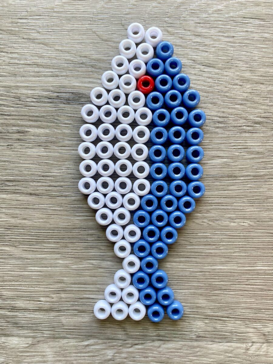 A fish shape made from white and blue beads with one red bead for an eye, arranged on a wooden surface.