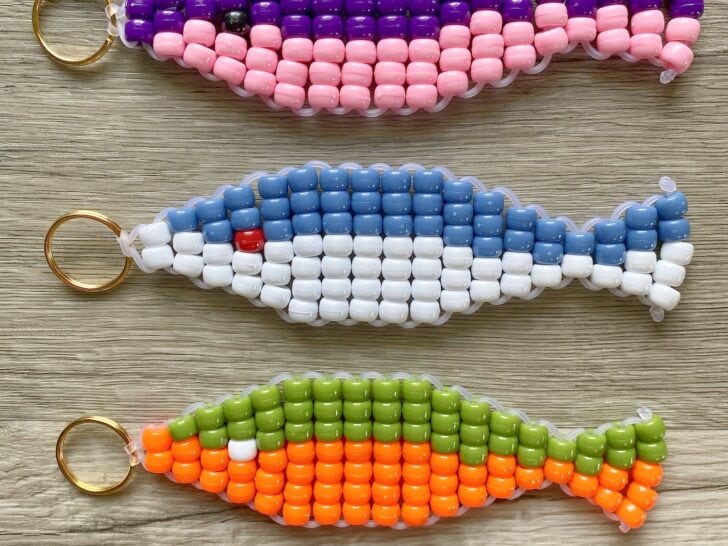 Three fish-shaped beaded keychains in purple-pink, blue-white, and green-orange patterns displayed on a wooden surface.