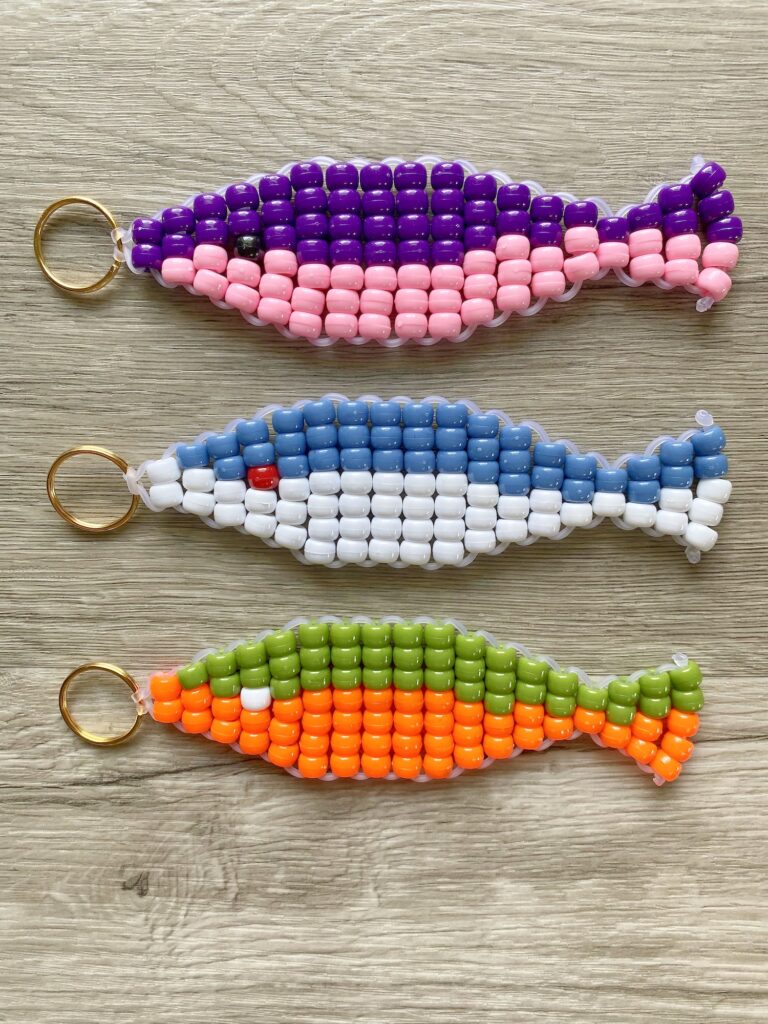Three fish-shaped beaded keychains in purple-pink, blue-white, and green-orange patterns displayed on a wooden surface.