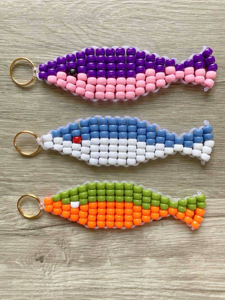 Three fish-shaped beaded keychains in purple-pink, blue-white, and green-orange patterns displayed on a wooden surface.