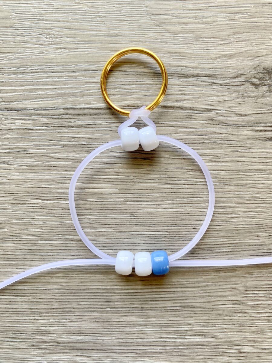 A gold keyring with a loop of clear cord, threaded with three white beads at the top and three beads (two white, one blue) at the bottom, placed on a wooden surface.