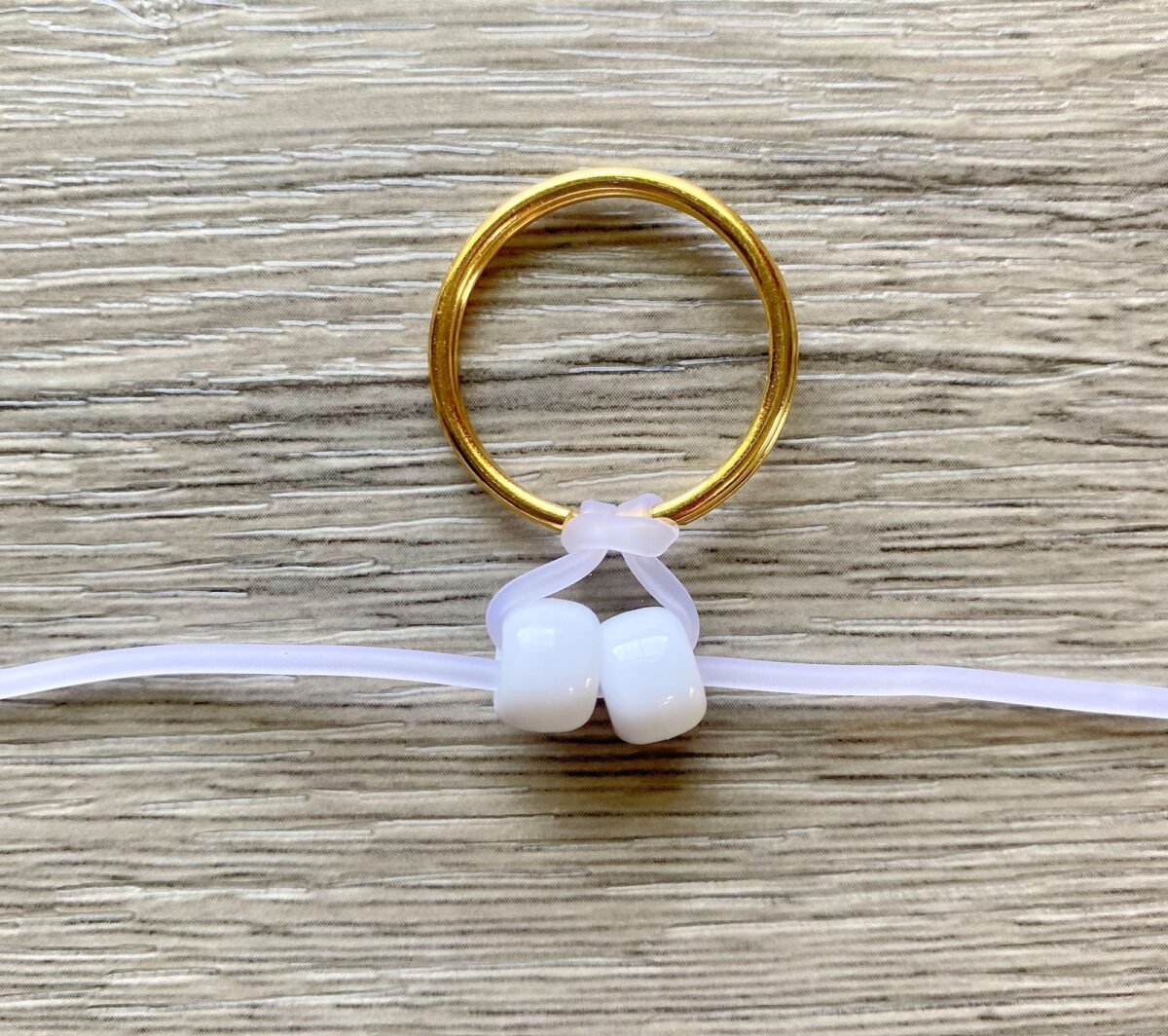 A gold ring with a white elastic strap threaded through it, secured by two white beads, on a wooden surface.
