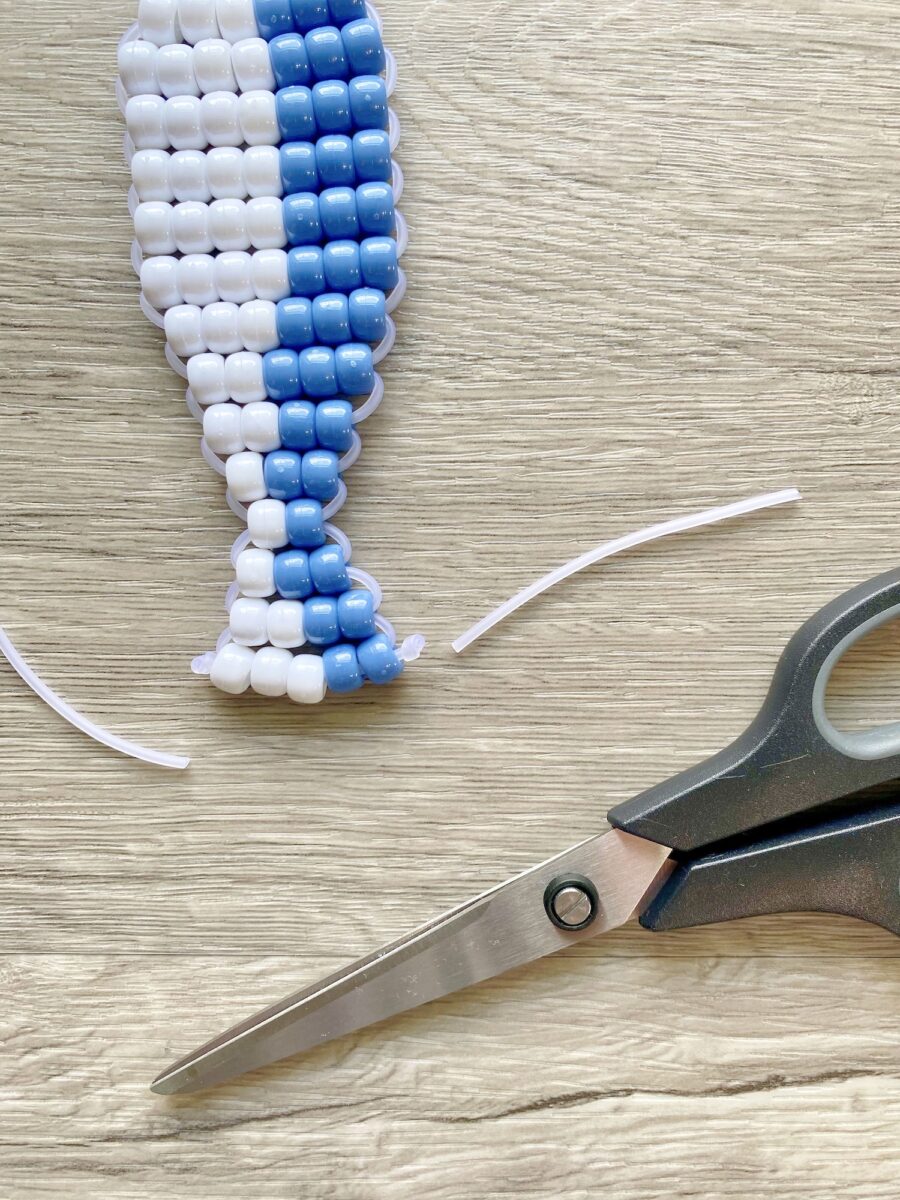 A pair of scissors and a beaded blue and white craft project with trimmed ends of string on a wooden surface.