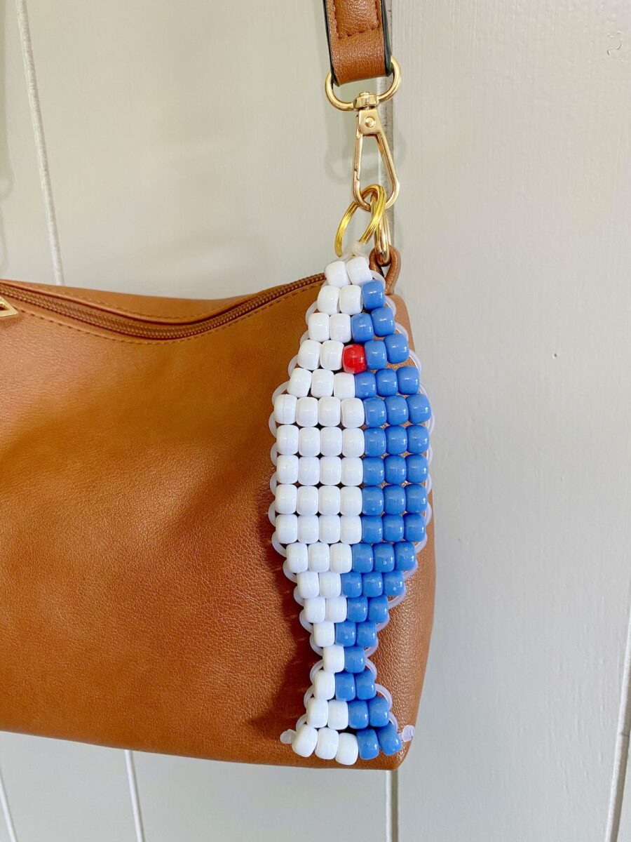 A keychain made of blue, white, and red beads in the shape of a fish is attached to a brown leather bag.