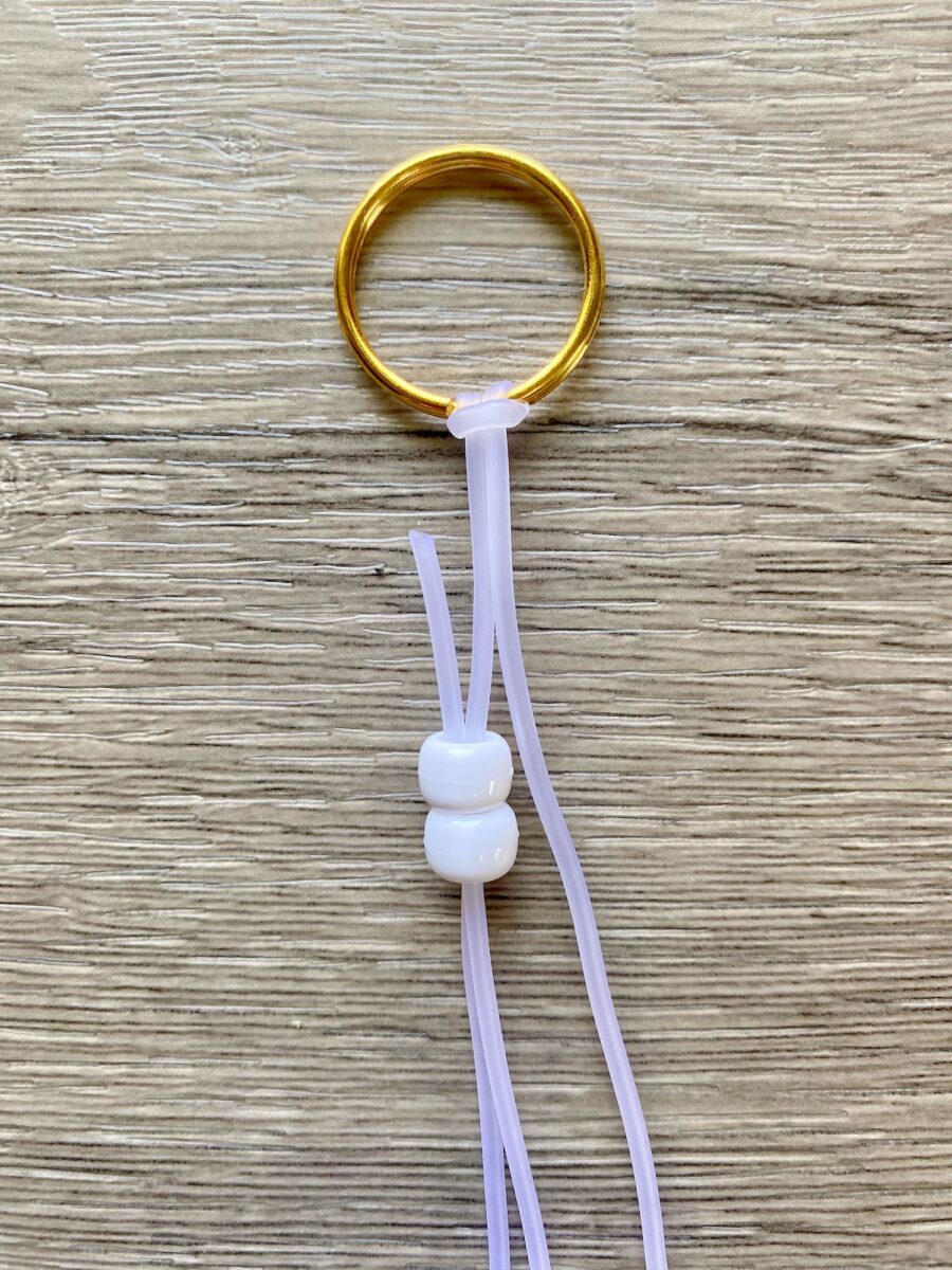 A gold key ring with two pale purple cords, each threaded through two white beads, lies on a wooden surface.