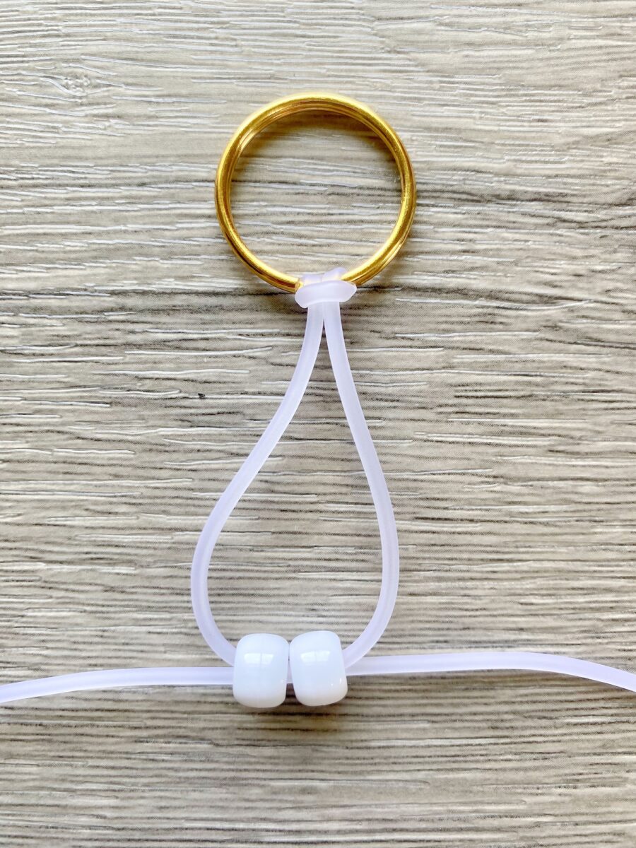 A gold keyring with a white elastic cord looped through it, holding two white beads, is displayed on a light wood surface.