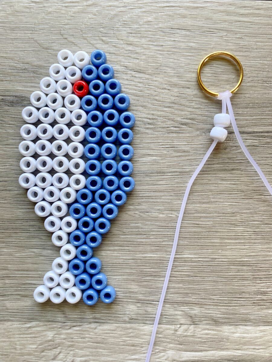 Perler bead fish design in blue, white, and one red bead beside a key ring with a bead on a white cord, on a wooden surface.
